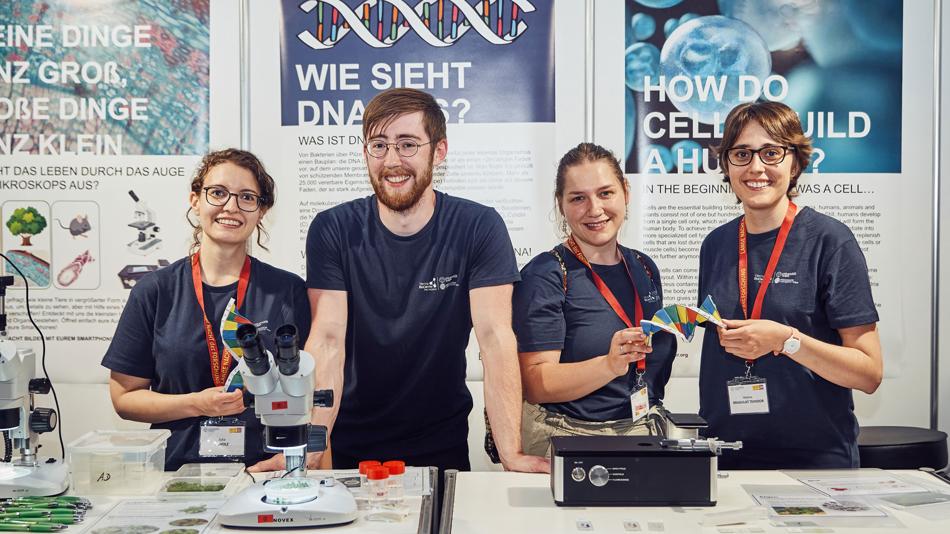 Infostand "Wie sieht DNA aus?" mit 4 jungen Molekularbiolog*innen mit Mikroskop