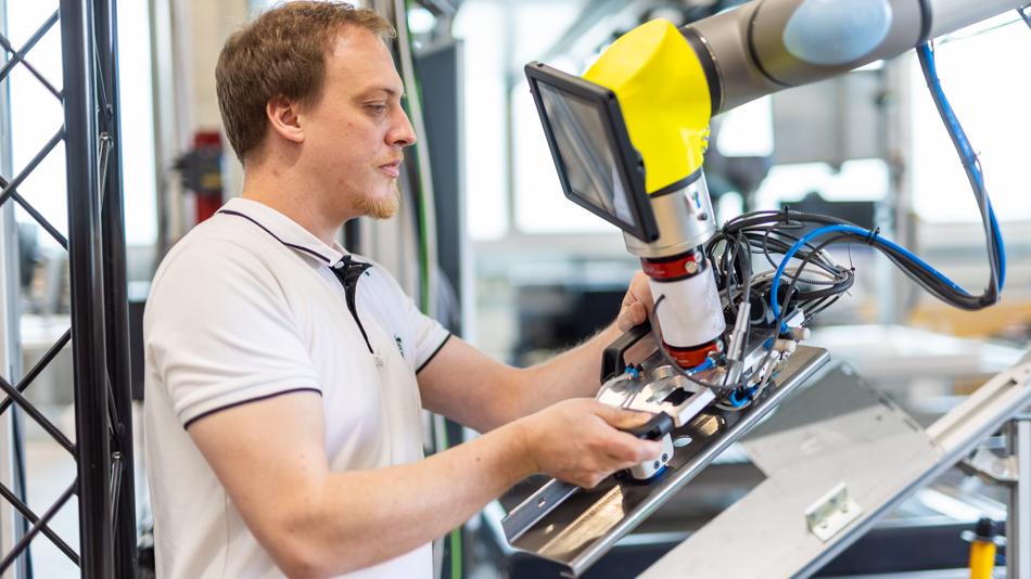 Ein Mitarbeiter in einem Industrie-Labor bedient einen kollaborativen Roboterarm (Cobot) mit Bedienpanel und montiert dabei ein Bauteil an einer Anlage.