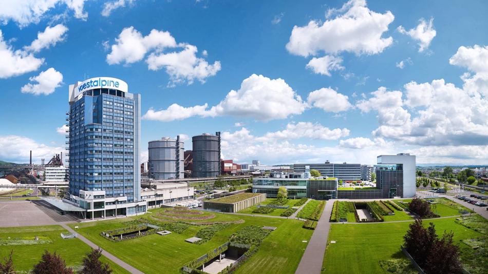 Panorama des voestalpine Standorts in Linz mit Bürogebäude, Industrieanlagen und weitläufigen Grünflächen unter blauem Himmel.