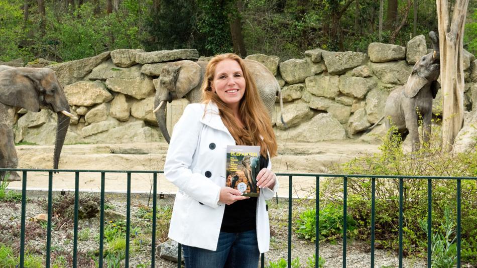 Angela Stöger hält ihr Buch "Elefanten" in den Händen. Hinter ihr sind echte Elefanten im Zoo zu sehen.