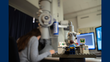 Im Hintergrund unscharf Person an einem großen Mikroskop, im Vordergrund Mikroskop aus Lego mit Lego-Figur