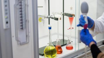 Mehrere gläserne Laborzylinder mit farbigem Inhalt, zwei Hände in blauen Handschuhen, welche ein Laborgerät halten.