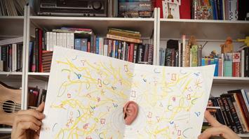 hochgehaltene Soundmapping-Zeichnung mit in der Mitte durchgestecktem Ohr vor Bücherregal