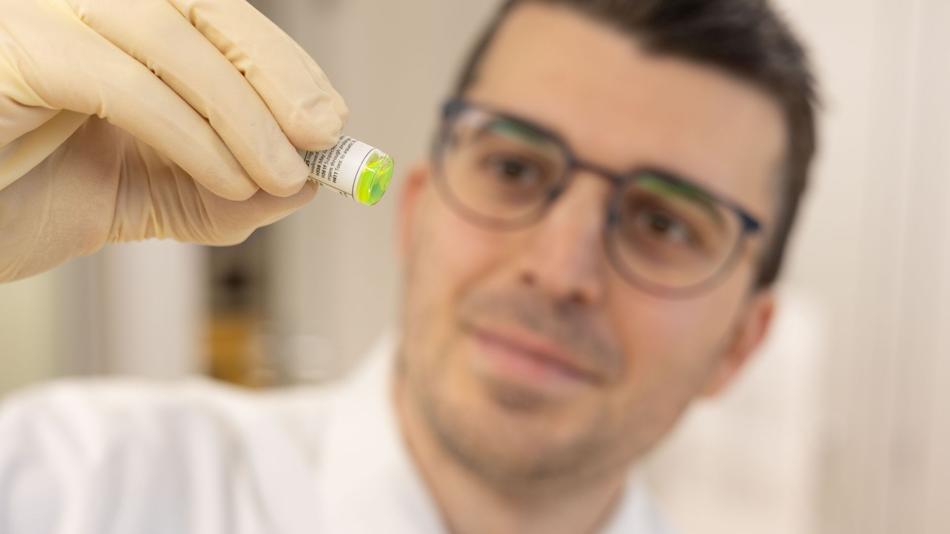 Eine behandschuhte Hand hält eine Probe mit leuchtend grünem Inhalt; im Hintergrund ist eine Person unscharf zu sehen.