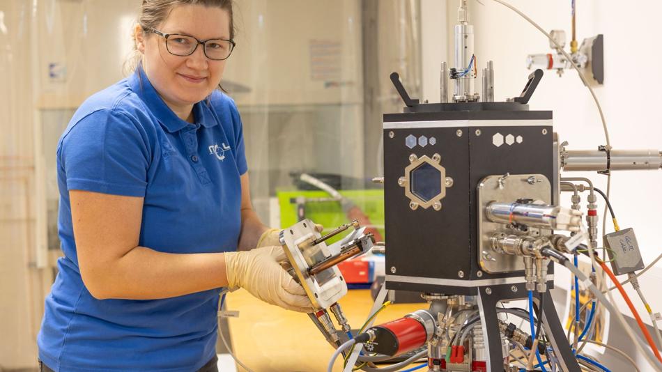 Eine Frau im blauen Poloshirt und mit Schutzhandschuhen hält einen Probenhalter neben einem Gerät in einem Labor der Mikroelektronik.