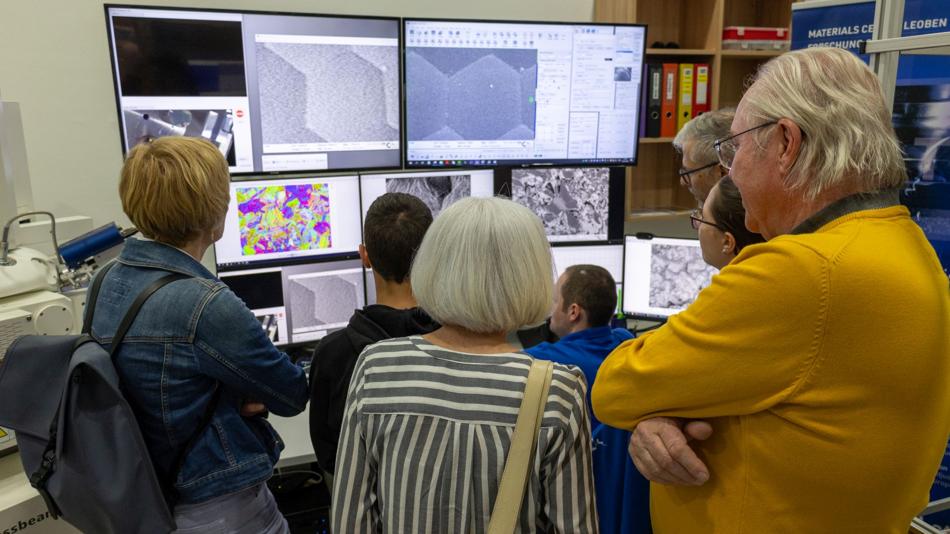 Mehrere Personen stehen im Labor vor einer Monitorwand mit Mikroskopie- und Materialbildern und verfolgen die Auswertung.