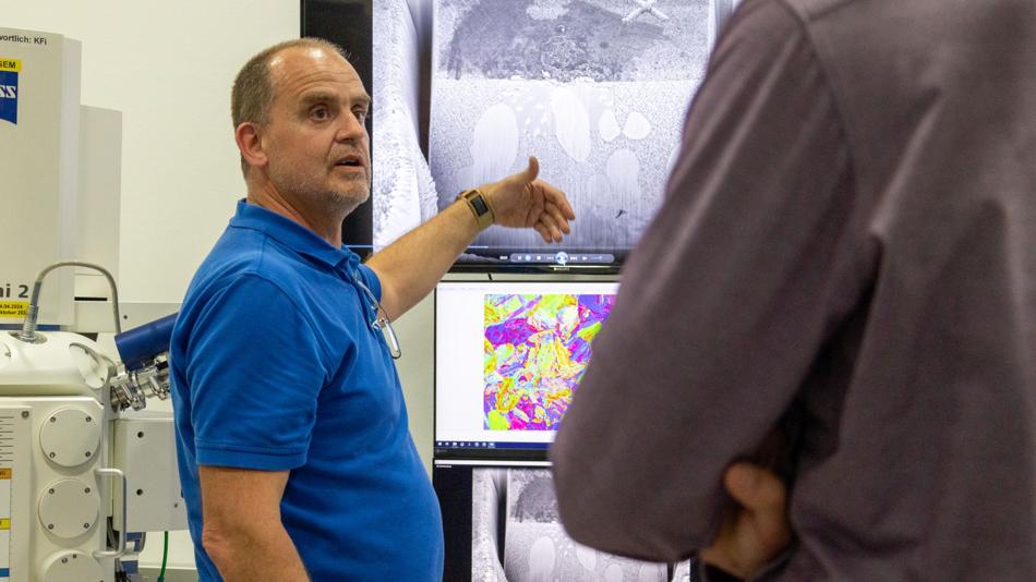 Ein Mann im blauen Poloshirt zeigt auf große Monitore mit mikroskopischen Aufnahmen, während eine weitere Person im Vordergrund zuhört.