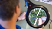 Durch eine Lupe sind Bauteile auf einer grünen Leiterplatte zu sehen, während eine Person im Vordergrund unscharf arbeitet.