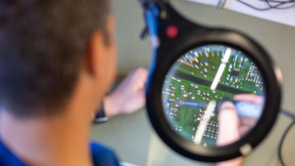 Durch eine Lupe sind Bauteile auf einer grünen Leiterplatte zu sehen, während eine Person im Vordergrund unscharf arbeitet.