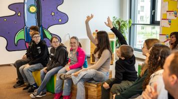 Kinder/Jugendliche sitzen auf Hocker und zeigen auf