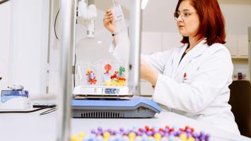 Eine Forscherin betrachtet konzentriert ein Experiment in einer Petrischale, während im Vordergrund farbenfrohe, stilisierte Molekülmodel