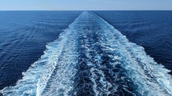 Blick vom Heck eines Schiffes auf das offene Meer, wobei eine lange, weiße Gischtspur in der Mitte zurückbleibt und sich bis zum Horizont erstreckt. Der Himmel ist klar und blau, ebenso wie das umgebende Wasser.