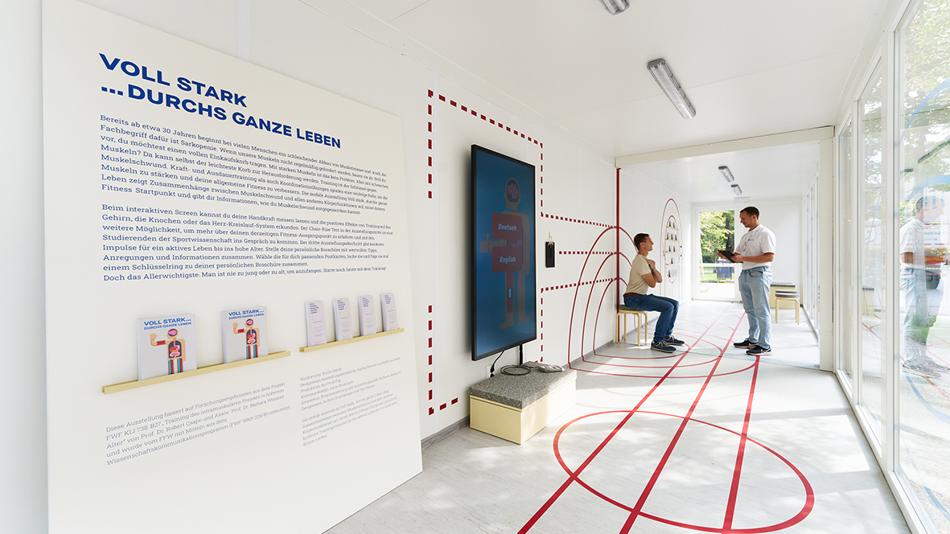 Durchführung eines Aufsteh Fitnesstests. Zwei Männer in einem Lichtdurchfluteten Ausstellungcontainer, einer hält ein Klemmbrett, der andere sitzt auf einem Hocker und ist bereit, aufzustehen.