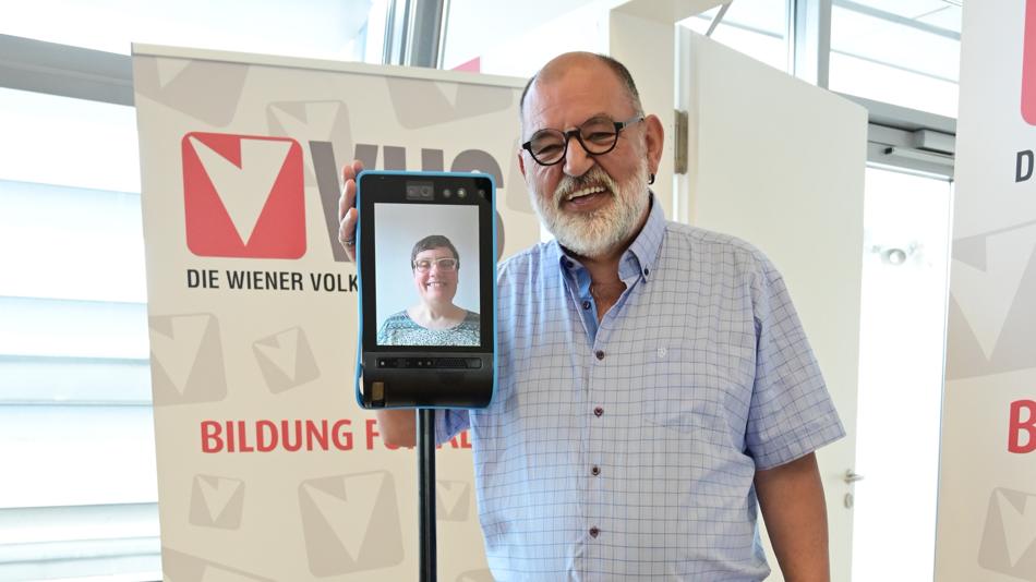 Zwei Personen stehen nebeneinander. Eine Person ist vor Ort, die andere nutzt einen Avatar. Im Hintergrund sieht man VHS Plakate.