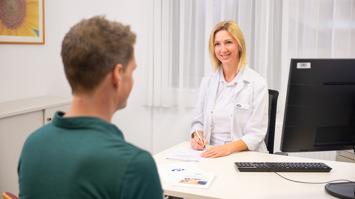 Darstellung eines Aufnahmegesprächs. Ein Mann wird von einer Ärztin aufgenommen. Beide sitzen sich an einem Schreibtisch gegenüber.