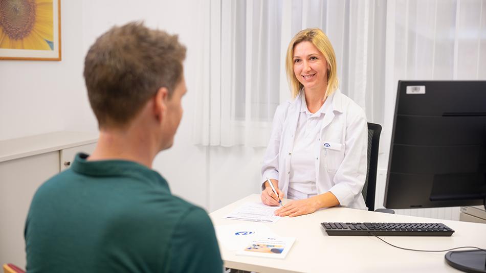 Darstellung eines Aufnahmegesprächs. Ein Mann wird von einer Ärztin aufgenommen. Beide sitzen sich an einem Schreibtisch gegenüber.