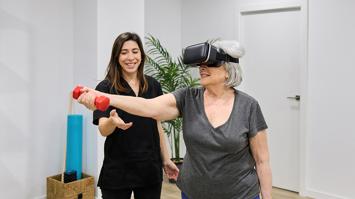Eine Rehabilitandin führt mit der Unterstützung einer Physiotherapeutin eine VR-gestütze Therapie durch und hat eine Hantel in der rechten Hand.