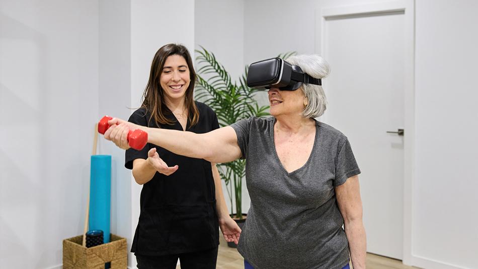 Eine Rehabilitandin führt mit der Unterstützung einer Physiotherapeutin eine VR-gestütze Therapie durch und hat eine Hantel in der rechten Hand.