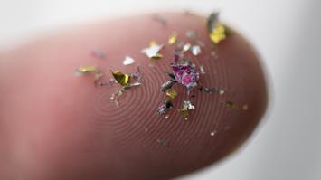 Nahaufnahme eines Fingers mit zahlreichen kleinen, bunten Mikroplastik-Partikeln darauf