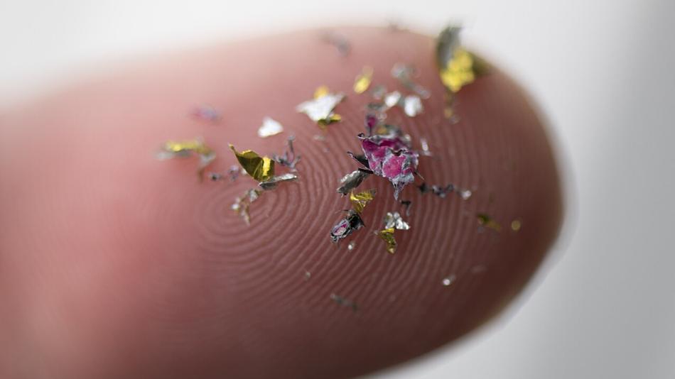 Nahaufnahme eines Fingers mit zahlreichen kleinen, bunten Mikroplastik-Partikeln darauf