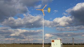 Kleine Windkraftanlage auf einem Mast neben einem Technikcontainer unter bewölktem Himmel