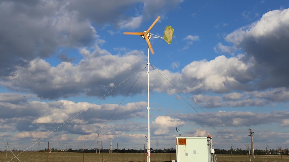 Kleine Windkraftanlage auf einem Mast neben einem Technikcontainer unter bewölktem Himmel