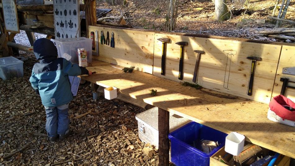Ein Kind steht an einer hölzernen Werkbank im Wald. An der Wand hängen Hämmer und andere Werkzeuge, während das Kind mit Naturmaterialien arbeitet.