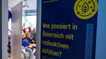 Plakat mit dem Namen der Station "Was passiert in Österreich mit radioaktiven Abfällen?". Im Hintergrund Besucherinnen und Besucher der Langen Nacht der Forschung