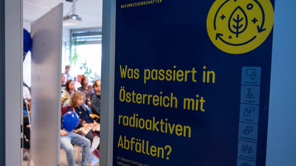 Plakat mit dem Namen der Station "Was passiert in Österreich mit radioaktiven Abfällen?". Im Hintergrund Besucherinnen und Besucher der Langen Nacht der Forschung