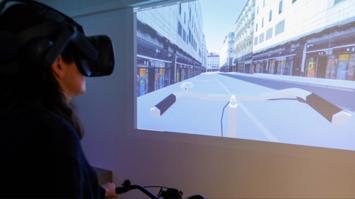 Eine Person mit VR-Brille erlebt eine virtuelle Fahrradszene auf großer Leinwand