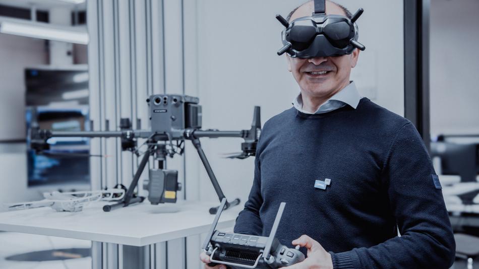 Ein Mann mit Virtual-Reality-Brille hält eine Drohnensteuerung; hinter ihm steht eine professionelle Drohne auf einem Tisch in einem Labor oder Technikraum.