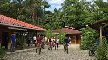 Mehrere Personen auf Rädern oder stehend auf einem Schotterboden, links ein Haus und im Hintergrund tropischer Regenwald mit Palmen und Bäumen