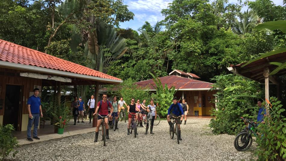 Mehrere Personen auf Rädern oder stehend auf einem Schotterboden, links ein Haus und im Hintergrund tropischer Regenwald mit Palmen und Bäumen