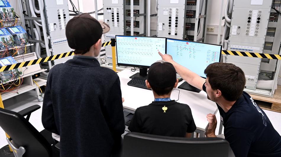 Ein Mitarbeiter von Sprecher Automation erklärt zwei Kindern vor Bildschirmen, wie das Stromnetz funktioniert.