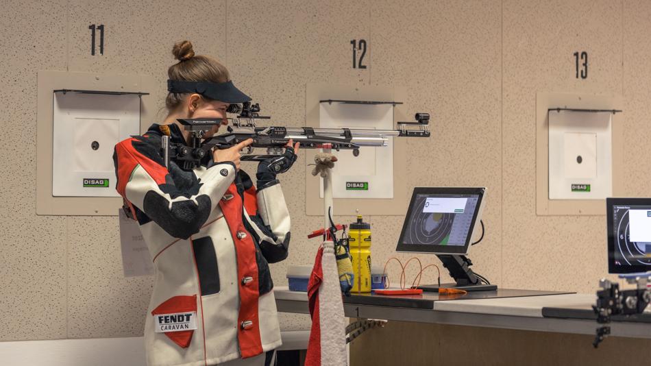 Person in Schießsport-Ausrüstung zielt mit einem Sportgewehr in einer Indoor-Schießanlage neben digitaler Zielanzeige.