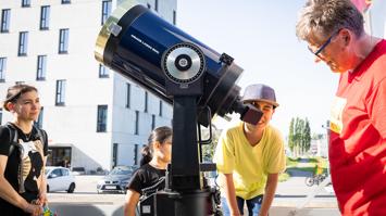 Kinder und ein Erwachsener betrachten durch ein großes Teleskop im Freien.