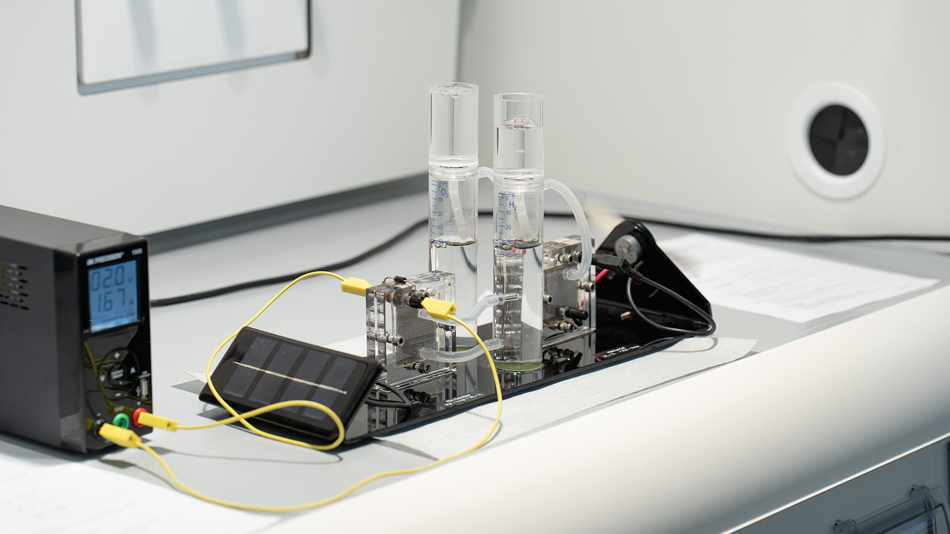 Ein Labor-Versuchsaufbau zur Elektrolyse von Wasser auf einer weißen Arbeitsfläche. Links steht ein digitales Netzgerät, das über gelbe Kabel mit einem PEM-Elektrolyseur und einer kleinen Solarzelle verbunden ist. Im Zentrum stehen zwei transparente, mit