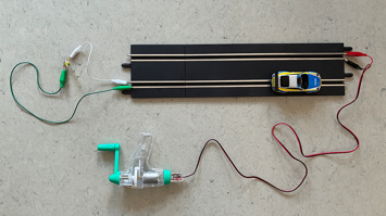 Handkurbel‑Generator ist per Kabel mit einem Carrera‑Bahnstück verbunden, darauf steht ein kleines Polizeiauto.