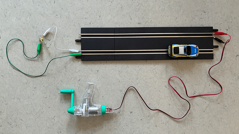 Handkurbel‑Generator ist per Kabel mit einem Carrera‑Bahnstück verbunden, darauf steht ein kleines Polizeiauto.