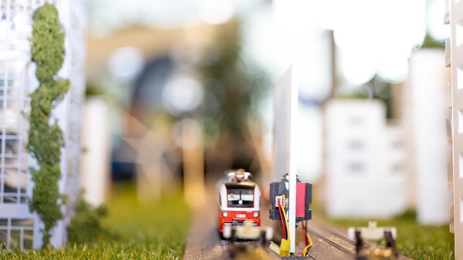 Nahaufnahme einer Modellbahn-Szenerie: ein roter ÖBB-Zug fährt auf Gleisen durch eine Miniaturstadt mit Gebäuden, Grasflächen und technischen Komponenten.
